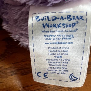 Puede incluir: Primer plano de una etiqueta de Build-A-Bear Workshop, con el nombre de la marca en azul. La etiqueta incluye el texto "Where Best Friends Are Made" y "Stuffed with Hugs and Good Wishes". La etiqueta tambi&eacute;n indica el origen del producto: China.