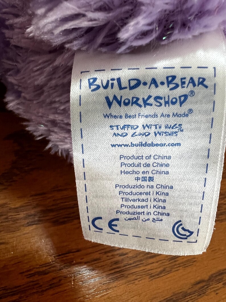 Puede incluir: Primer plano de una etiqueta de Build-A-Bear Workshop, con el nombre de la marca en azul. La etiqueta incluye el texto "Where Best Friends Are Made" y "Stuffed with Hugs and Good Wishes". La etiqueta tambi&eacute;n indica el origen del producto: China.