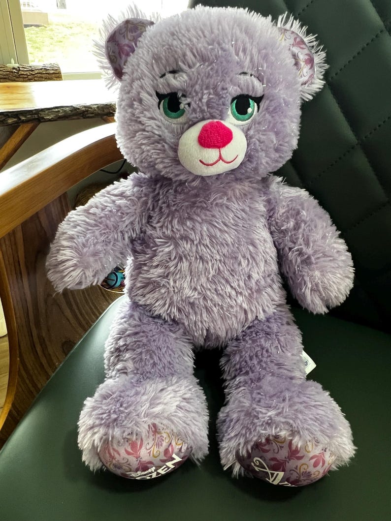 Puede incluir: Un oso de peluche lavanda con una nariz rosa, ojos verdes y un hocico blanco. El oso tiene una textura suave y esponjosa y detalles decorativos en sus patas. El oso est&aacute; sentado en una silla verde oscuro.
