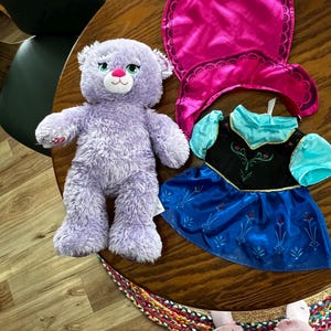 Puede incluir: Un oso de peluche lavanda con nariz rosa y ojos verdes est&aacute; junto a una capa rosa y un vestido azul y negro con mangas turquesas. Los art&iacute;culos est&aacute;n sobre una mesa de madera.