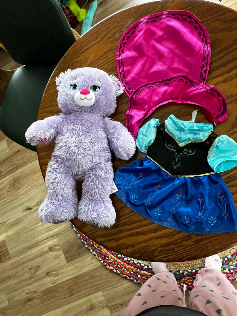 Puede incluir: Un oso de peluche lavanda con nariz rosa y ojos verdes est&aacute; junto a una capa rosa y un vestido azul y negro con mangas turquesas. Los art&iacute;culos est&aacute;n sobre una mesa de madera.
