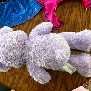 Puede incluir: Un oso de peluche lavanda con una etiqueta, acostado sobre una superficie de madera. El oso tiene una textura esponjosa. Se ven piezas de disfraz, incluyendo una falda azul con detalles florales y una diadema rosa.