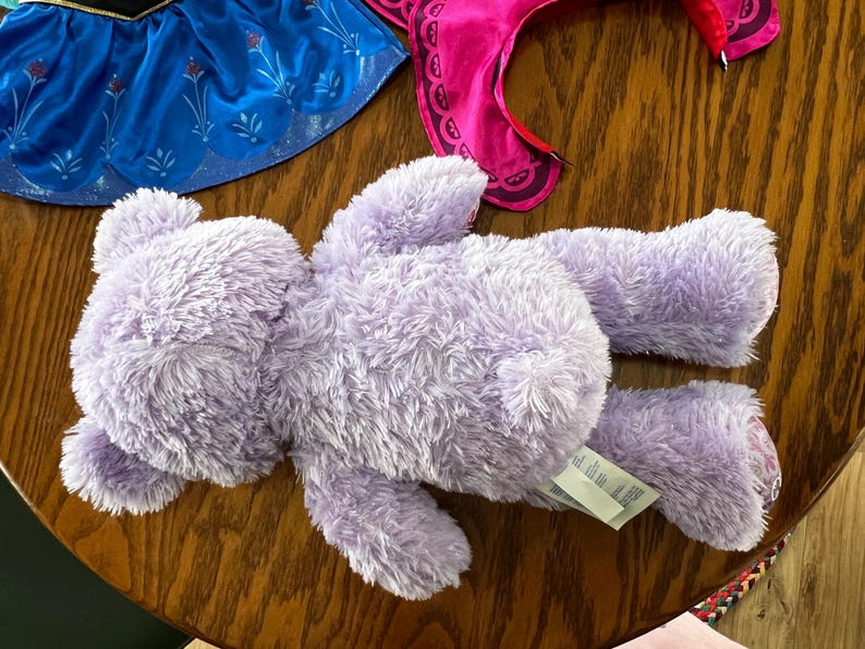 Puede incluir: Un oso de peluche lavanda con una etiqueta, acostado sobre una superficie de madera. El oso tiene una textura esponjosa. Se ven piezas de disfraz, incluyendo una falda azul con detalles florales y una diadema rosa.