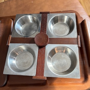 May include: A square wooden serving tray with four stainless steel bowls. The tray has a dark brown wood finish and the bowls are polished.