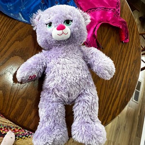 Puede incluir: Un oso de peluche lavanda con hocico blanco y nariz rosa. El oso tiene ojos verdes y est&aacute; sobre una mesa de madera. Otros art&iacute;culos coloridos est&aacute;n en el fondo, incluyendo un disfraz azul y rosa.