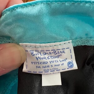 Puede incluir: Primer plano de una etiqueta de un producto Build-A-Bear Workshop. La etiqueta blanca tiene texto azul que dice "BUILD-A-BEAR WORKSHOP STITCHED WITH LOVE FOR AGES 3 AND UP Made in China."