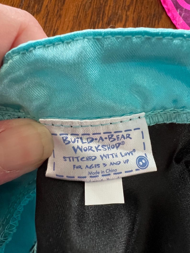 Puede incluir: Primer plano de una etiqueta de un producto Build-A-Bear Workshop. La etiqueta blanca tiene texto azul que dice "BUILD-A-BEAR WORKSHOP STITCHED WITH LOVE FOR AGES 3 AND UP Made in China."