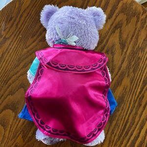 Puede incluir: Un oso de peluche morado con una capa rosa con un borde decorativo. El oso lleva un atuendo azul y turquesa. Un lazo blanco est&aacute; sujeto a la parte posterior de la cabeza del oso.