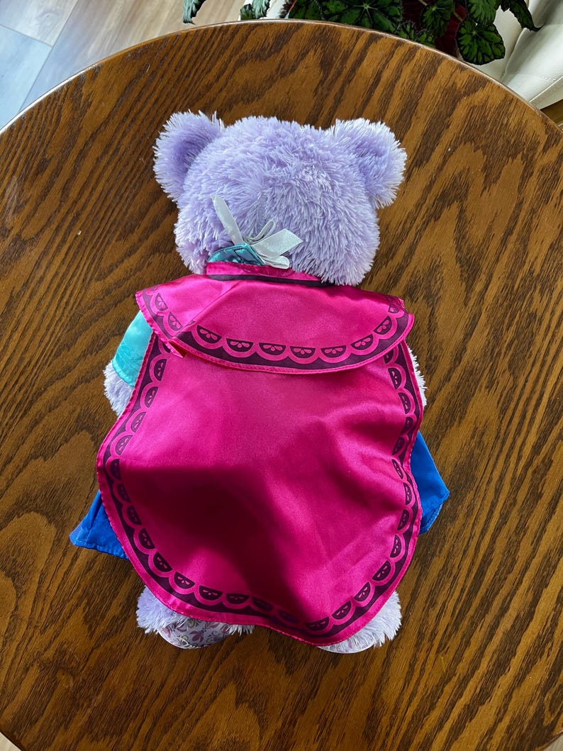 Puede incluir: Un oso de peluche morado con una capa rosa con un borde decorativo. El oso lleva un atuendo azul y turquesa. Un lazo blanco est&aacute; sujeto a la parte posterior de la cabeza del oso.
