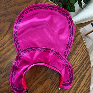 Puede incluir: Un conjunto de protecci&oacute;n solar rosa brillante que incluye una visera y un gorro a juego, ambos con un borde festoneado de color p&uacute;rpura oscuro. La visera tiene una visera curva y un interior rojo.