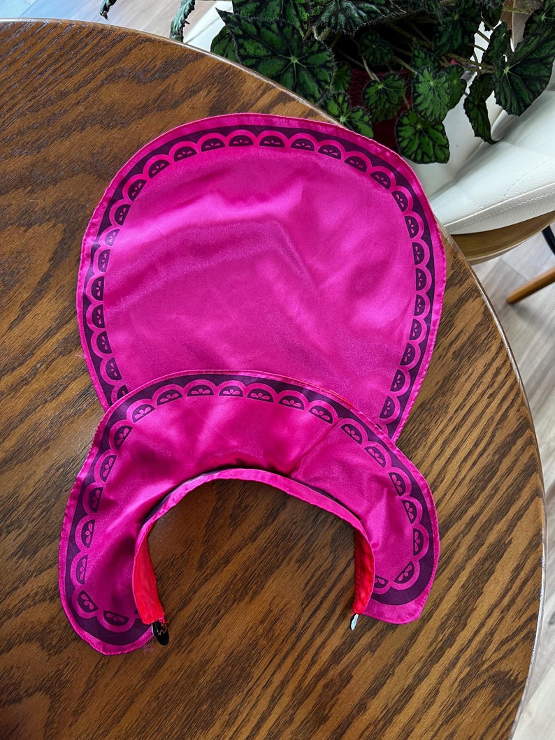 Puede incluir: Un conjunto de protecci&oacute;n solar rosa brillante que incluye una visera y un gorro a juego, ambos con un borde festoneado de color p&uacute;rpura oscuro. La visera tiene una visera curva y un interior rojo.
