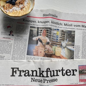 Op de afbeelding: Een kom yoghurt met granola, bosbessen, bananenschijfjes en appels. De kom staat op een krant met Duitse tekst en krantenkoppen, waaronder "Frankfurter Neue Presse".
