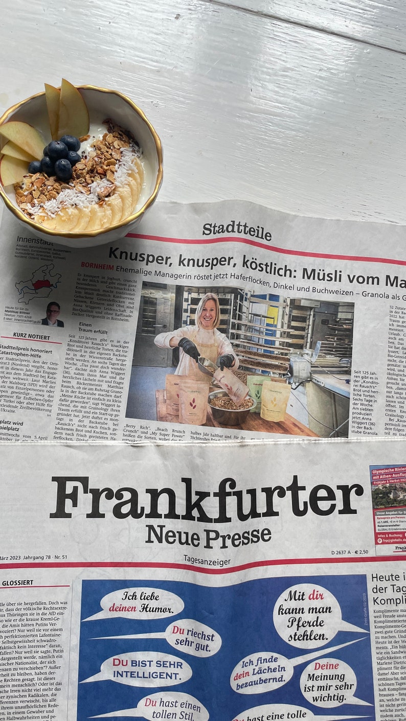 Op de afbeelding: Een kom yoghurt met granola, bosbessen, bananenschijfjes en appels. De kom staat op een krant met Duitse tekst en krantenkoppen, waaronder "Frankfurter Neue Presse".