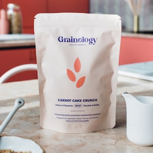 Op de afbeelding: Een witte zak Grainology 'Carrot Cake Crunch' granola. De zak heeft de merknaam in blauw, een oranje ontwerp en productdetails. Een witte kan en een kom met granola staan op de voorgrond.