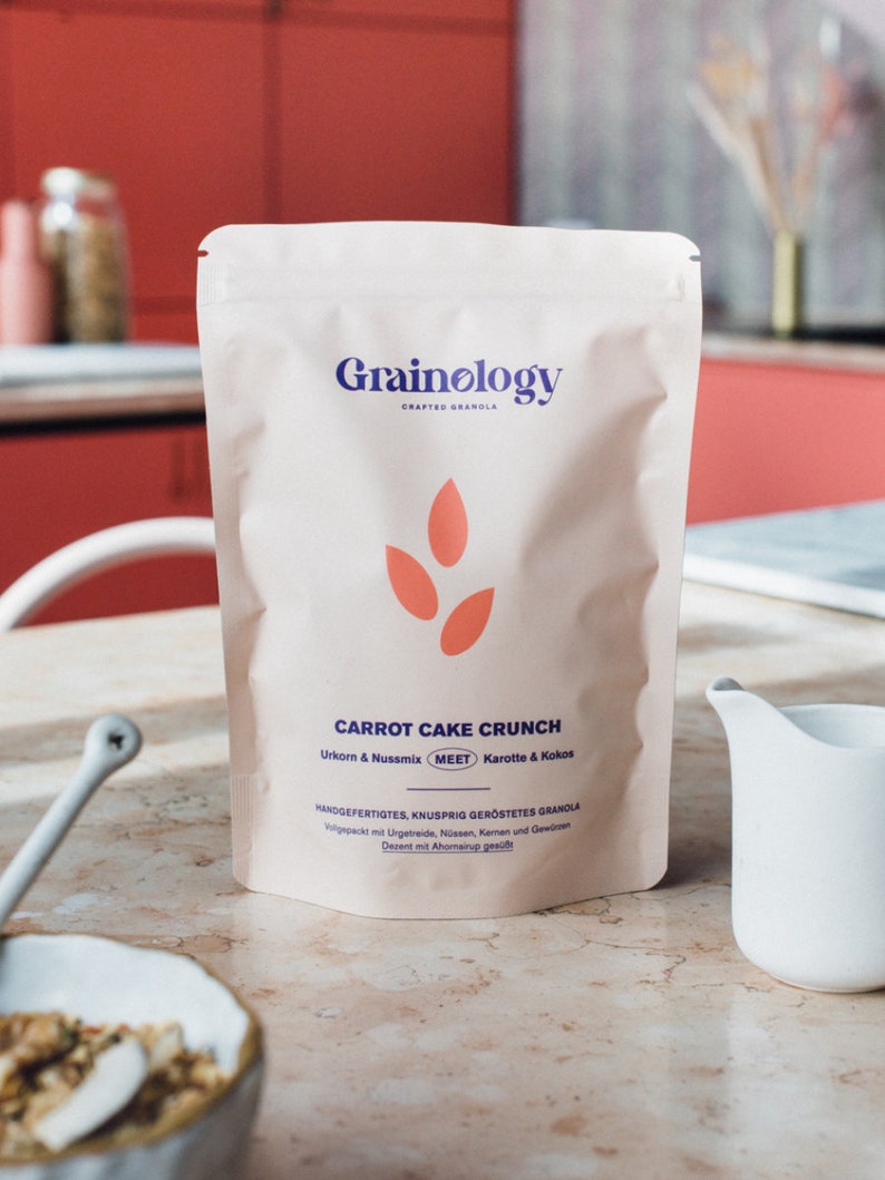 Op de afbeelding: Een witte zak Grainology 'Carrot Cake Crunch' granola. De zak heeft de merknaam in blauw, een oranje ontwerp en productdetails. Een witte kan en een kom met granola staan op de voorgrond.