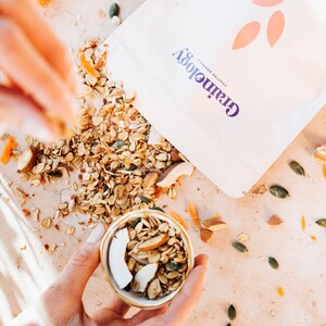 Op de afbeelding: Een close-up van een kom gevuld met granola, kokosvlokken en amandelen. De granola is verspreid over een lichtgekleurd oppervlak, met een zak Grainology granola op de achtergrond. Wortelen en pompoenpitten zijn ook zichtbaar.