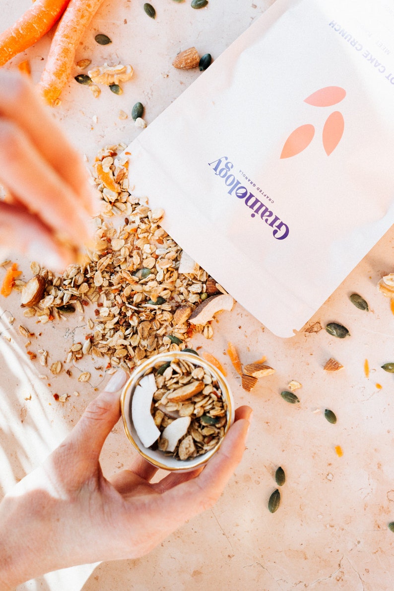 Op de afbeelding: Een close-up van een kom gevuld met granola, kokosvlokken en amandelen. De granola is verspreid over een lichtgekleurd oppervlak, met een zak Grainology granola op de achtergrond. Wortelen en pompoenpitten zijn ook zichtbaar.