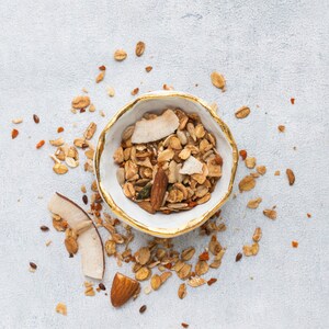 Op de afbeelding: Een witte kom met een gouden rand gevuld met granola, kokosvlokken en amandelen. Granola is verspreid rond de kom op een lichtgrijze ondergrond. De granola lijkt een mix van haver, noten en gedroogd fruit te zijn.