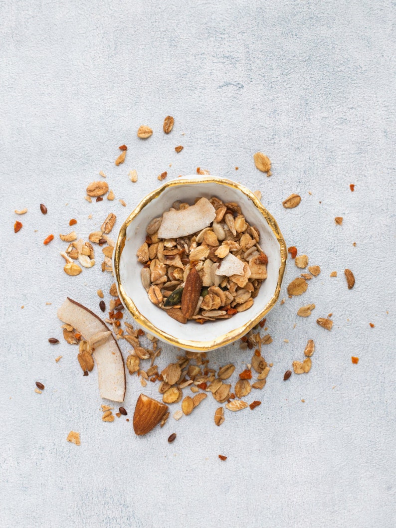 Op de afbeelding: Een witte kom met een gouden rand gevuld met granola, kokosvlokken en amandelen. Granola is verspreid rond de kom op een lichtgrijze ondergrond. De granola lijkt een mix van haver, noten en gedroogd fruit te zijn.
