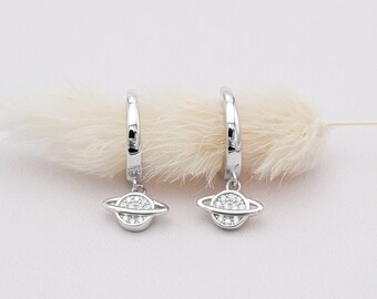 May include: A pair of silver hoop earrings with a planet charm. Each charm features a ring around a circular center, both encrusted with small, clear stones. The earrings are displayed against a soft, white background with a feathery accent.