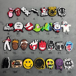 May include: A set of 29 colourful shoe charms featuring various popular music and pop culture icons. The charms include logos, characters, and phrases like "WAP", "AC/DC", "Metallica", "Nirvana", "Rock Till The End", "The Beatles", "Ghostbusters", "Snoop Dogg", "Kiss", and "Rock".