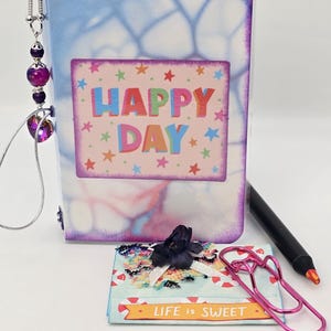 May include: A clear, decorative journal with a watercolor-style cover and the words "HAPPY DAY" on the front. A black pen, a pink paperclip, and a small gift with a black bow are also visible.
