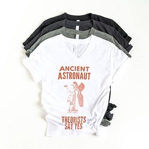May include: A stack of v-neck t-shirts in white, gray, and black. The white shirt features the text "ANCIENT ASTRONAUT" above an illustration of a figure with wings, and the text "THEORISTS SAY YES" below.