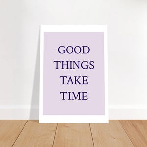 Könnte beinhalten: Gerahmter Druck mit den Worten „GOOD THINGS TAKE TIME“ in dunkelblauen Buchstaben auf helllila Hintergrund. Der Druck lehnt an einer weißen Wand auf einem Holzboden.