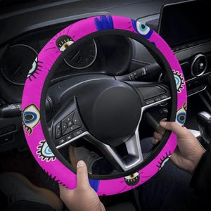 May include: A pink steering wheel cover featuring a repeating pattern of blue and white eye designs. The cover is fitted on a black steering wheel inside a car. The cover is held by a person's hands.