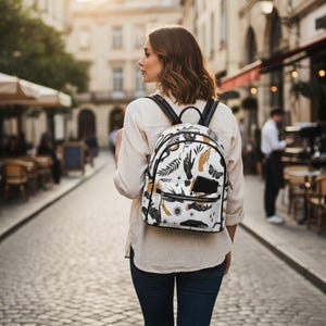 Könnte beinhalten: Ein weißer Rucksack mit einem schwarzen und goldenen Himmelsdesign, einschließlich Händen, Schmetterlingen und Sternen. Der Rucksack hat schwarze Träger und eine Vordertasche. Die Person geht eine Kopfsteinpflasterstraße entlang.