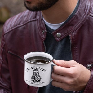May include: White enamel mug with a black rim and handle. The mug features the text "PLANT DADDY" above an illustration of a bearded man with glasses and a leafy beard. Below the illustration is the text "THE ONLY MAN WHO CAN GROW A BEARD AND A JUNGLE."