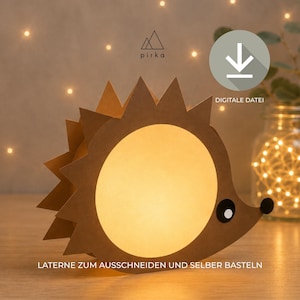 Può includere: Una lanterna a forma di riccio in carta marrone con un grande centro circolare luminoso, un occhio nero e un naso nero. Il design presenta il testo 'pirka' con un logo a forma di montagna, un'icona di download con 'DIGITALE DATEI' e 'LATERNE ZUM AUSSCHNEIDEN UND SELBER BASTELN'.