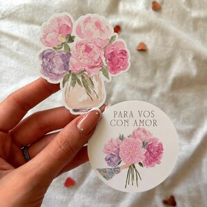 Puede incluir: Una pegatina troquelada con una ilustración en acuarela de peonías rosas y moradas en un jarrón, junto a una pegatina circular con un ramo de peonías y el texto "PARA VOS CON AMOR" impreso en una tipografía con remates.