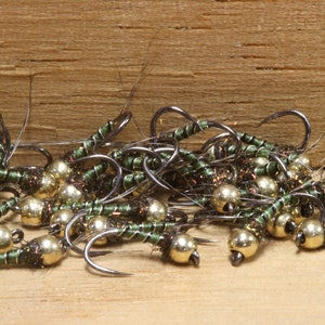 Può includere: Primo piano di un gruppo di esche da pesca a mosca verdi e dorate. Le esche sono legate con filo verde e hanno una testa di perline dorate.