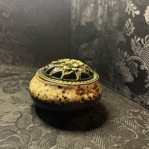 May include: A black and brown ceramic incense burner with a gold metal floral design on the lid.