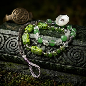 Puede incluir: Conjunto de joyas de cuentas verdes y transparentes, con cierre de botón. Las joyas se exhiben sobre una superficie de piedra tallada, con un anillo dorado en el fondo. Las cuentas tienen diseños florales.