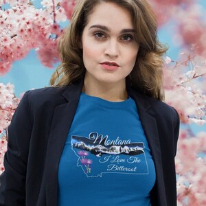 May include: A woman wearing a blue t-shirt with a graphic of the state of Montana and the text "Montana I Love the Bitterroot". She is wearing a black blazer and black pants. The background is a blurred image of pink cherry blossoms.