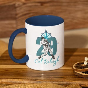 May include: White ceramic mug with a navy blue interior and handle. The mug features a baseball player graphic with the number 29 and the name "Cal Raleigh". A compass rose design is above the player.