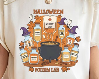 May include: Cream-colored t-shirt featuring a Halloween-themed graphic. The design includes a cauldron, potion bottles, witch hats, and the words "Halloween" and "Potion Lab." The shirt is a relaxed fit.