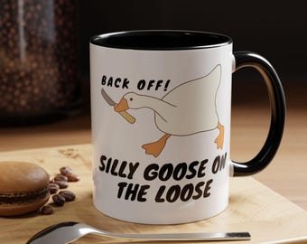 Könnte beinhalten: Weiße Keramiktasse mit schwarzem Rand und Henkel. Die Tasse zeigt eine Cartoon-Gans, die ein Messer hält, und den Text "Back Off! Silly Goose on the Loose".