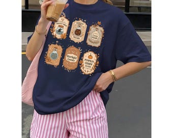 Puede incluir: Camiseta azul marino con seis diseños de etiquetas de pociones con textos como "Ghostly Brew" y "Pumpkin Charm Elixir". La camiseta se combina con unos shorts a rayas rosas y blancas. La persona sostiene una bebida en un vaso transparente.