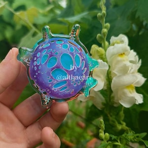 May include: A colorful, abstract glass art piece with a circular design. The piece features a vibrant mix of purple and turquoise hues, with a textured surface and a clear, green border. The text "by @althaçart" is visible on the piece.