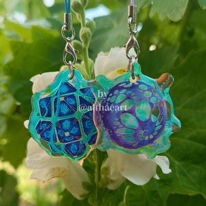 May include: Two colourful acrylic charms with silver-tone hardware. One charm is a blue geometric design, and the other is a purple and blue circular design. The charms are attached to blue lanyards. The text "by @althaeart" is visible.