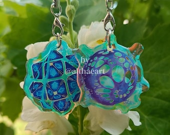 May include: Two colourful acrylic charms with silver-tone hardware. One charm is a blue geometric design, and the other is a purple and blue circular design. The charms are attached to blue lanyards. The text "by @althaeart" is visible.
