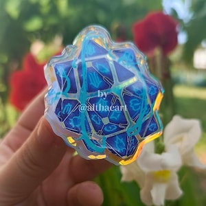 May include: A blue and gold geometric-shaped pin with a complex design. The pin features a series of blue shapes and lines, with the text "by / @althaeart" in the lower left corner. The pin is held in front of a blurred background of flowers.