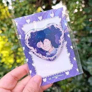 May include: A heart-shaped badge with an anime-style illustration of a person with dark blue hair and glasses. The badge is set against a purple background with a white lace border and heart pattern. Text on the card reads "by @althaeart".