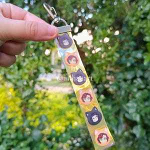 May include: A clear keychain strap with a silver clasp, featuring a repeating pattern of cartoon character faces in cat-shaped frames. The text "by @alhaart" is visible on the strap.