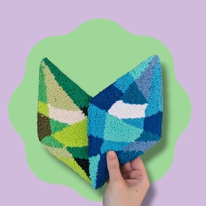 May include: Two geometric, tufted textile pieces. One is green with shades of olive, lime, and cream. The other is blue with shades of turquoise, navy, and white. The pieces are held against a green, wavy background on a purple backdrop.