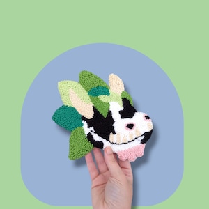 May include: A hand holds a green, black, and white cow-shaped rug made with yarn. The rug is a punch needle project.