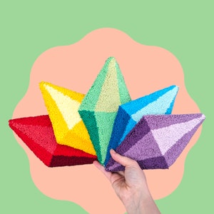 May include: A hand holds a fan of five colorful, geometric, diamond-shaped objects. The objects are red, yellow, green, blue, and purple. The background is a light green with a peach-colored shape.
